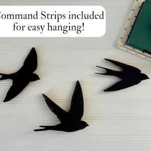 May include: Three black swallow wall art pieces in flight, mounted on a white paneled wall. The text "Command Strips included for easy hanging!" is in a white box above the birds. A decorative box with a flower is in the upper right corner.
