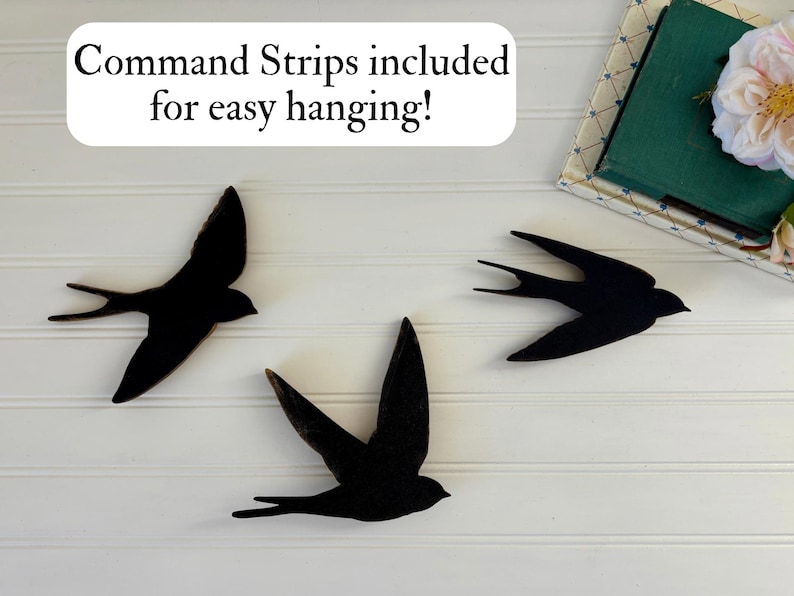 May include: Three black swallow wall art pieces in flight, mounted on a white paneled wall. The text "Command Strips included for easy hanging!" is in a white box above the birds. A decorative box with a flower is in the upper right corner.