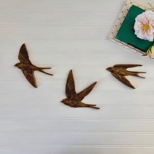 May include: Three bronze-colored swallow wall art pieces in flight, mounted on a white, horizontal-paneled surface. A green book with a floral arrangement and a white rose is in the upper right corner. The swallows have outstretched wings and detailed feathering.