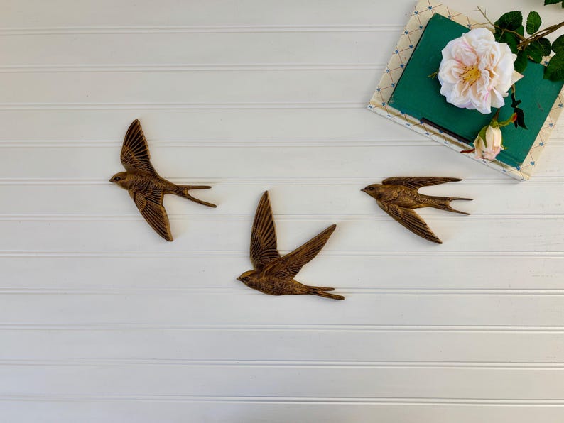 May include: Three bronze-colored swallow wall art pieces in flight, mounted on a white, horizontal-paneled surface. A green book with a floral arrangement and a white rose is in the upper right corner. The swallows have outstretched wings and detailed feathering.