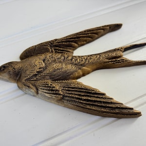 May include: A detailed, brown wooden bird sculpture with outstretched wings. The bird is carved with intricate patterns, showcasing the texture of the wood. The sculpture is set against a white background, highlighting the bird's form and design.