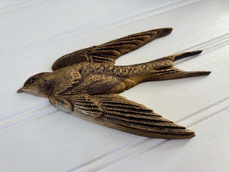 May include: A detailed, brown wooden bird sculpture with outstretched wings. The bird is carved with intricate patterns, showcasing the texture of the wood. The sculpture is set against a white background, highlighting the bird's form and design.