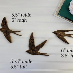 May include: Three bronze-colored metal swallow wall art pieces. The largest is 5.75 inches wide and 5.5 inches tall. The other two are 5.5 inches wide by 6 inches high, and 6 inches wide by 3.5 inches high. The birds are mounted on a white paneled wall.