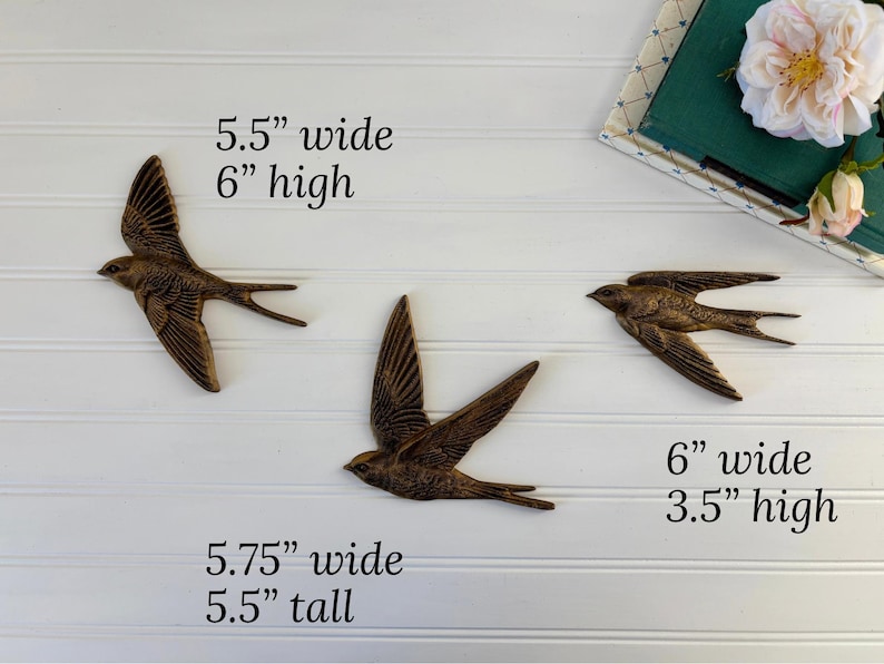 May include: Three bronze-colored metal swallow wall art pieces. The largest is 5.75 inches wide and 5.5 inches tall. The other two are 5.5 inches wide by 6 inches high, and 6 inches wide by 3.5 inches high. The birds are mounted on a white paneled wall.