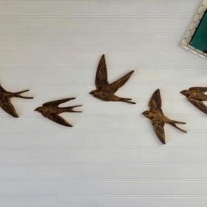 May include: Five bronze-colored swallows in flight are arranged on a white, horizontal-paneled wall. The birds have detailed wing and tail feathers. A book and flowers are in the upper right corner.