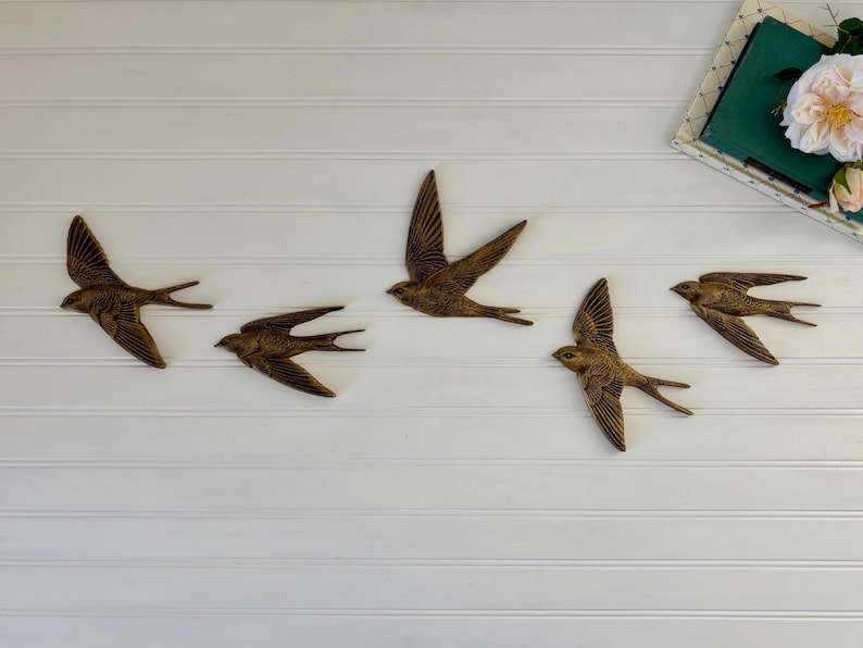 May include: Five bronze-colored swallows in flight are arranged on a white, horizontal-paneled wall. The birds have detailed wing and tail feathers. A book and flowers are in the upper right corner.
