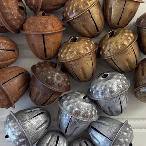 May include: A collection of decorative acorn-shaped bells in various metallic finishes. The bells are in shades of brown, bronze, and silver, with a textured surface and a slit for sound. These rustic bells are ideal for holiday decor.