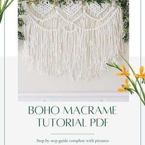 May include: A white macrame wall hanging with a geometric pattern and fringe. The hanging is decorated with green leaves and is displayed against a white wall. The text "BOHO MACRAME TUTORIAL PDF" is below the hanging, with the text "Step by step guide complete with pictures" below that.