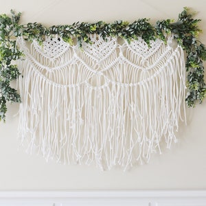 May include: White macrame wall hanging with a fringe and a garland of green leaves.