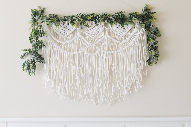 May include: White macrame wall hanging with a fringe and a garland of green leaves.