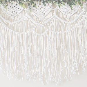 May include: White macrame wall hanging with a fringe border and a garland of green leaves at the top.