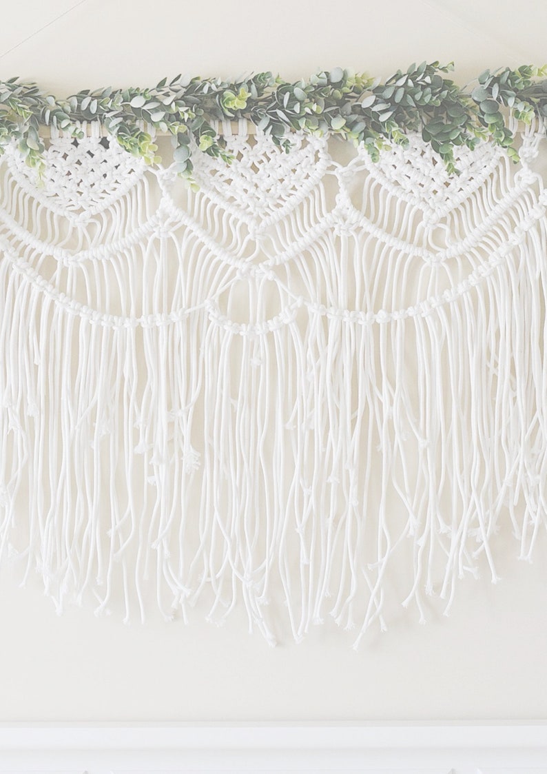 May include: White macrame wall hanging with a fringe border and a garland of green leaves at the top.