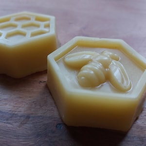 Puede incluir: Dos bloques de cera de abeja hexagonales, uno con un diseño de panal y el otro con un diseño de abeja.