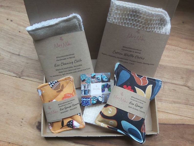 May include: A gift box filled with eco-friendly cleaning and bath products. The box contains a brown and white cotton waffle cloth, a brown and white eco cleaning cloth, a yellow and orange eco sponge scourer, and a black and blue eco sponge. The products are handmade in Wirral.