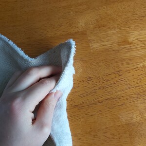 Puede incluir: Una mano sosteniendo un pa&ntilde;o de limpieza blanco y beige. El pa&ntilde;o est&aacute; doblado por la mitad y la mano lo sostiene por el borde doblado. El pa&ntilde;o est&aacute; sobre una superficie de madera.