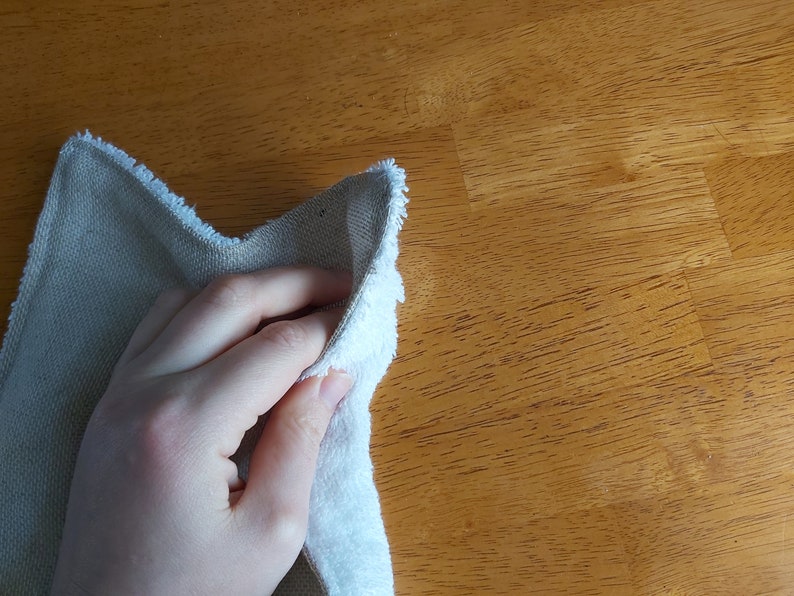 Puede incluir: Una mano sosteniendo un pa&ntilde;o de limpieza blanco y beige. El pa&ntilde;o est&aacute; doblado por la mitad y la mano lo sostiene por el borde doblado. El pa&ntilde;o est&aacute; sobre una superficie de madera.