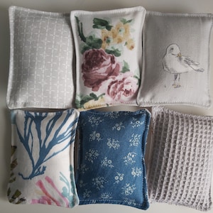 Puede incluir: Seis pequeños cojines rectangulares de tela con diferentes patrones. Los cojines están dispuestos en una fila, con el patrón floral en el centro. Los patrones incluyen un patrón geométrico blanco y gris, un patrón floral con flores rosas y amarillas, un patrón blanco y gris con un pájaro, un patrón blanco y azul con un diseño de coral, un patrón floral azul y blanco y un patrón de gofre gris.
