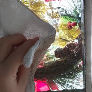 Puede incluir: Una mano sosteniendo un pa&ntilde;o de limpieza blanco y beige con una superficie texturizada. El pa&ntilde;o se est&aacute; utilizando para limpiar una ventana con un dise&ntilde;o de vidrio decorativo. La ventana est&aacute; decorada con una rama de pino verde y bayas rojas.