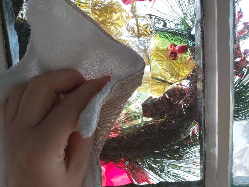 Puede incluir: Una mano sosteniendo un pa&ntilde;o de limpieza blanco y beige con una superficie texturizada. El pa&ntilde;o se est&aacute; utilizando para limpiar una ventana con un dise&ntilde;o de vidrio decorativo. La ventana est&aacute; decorada con una rama de pino verde y bayas rojas.
