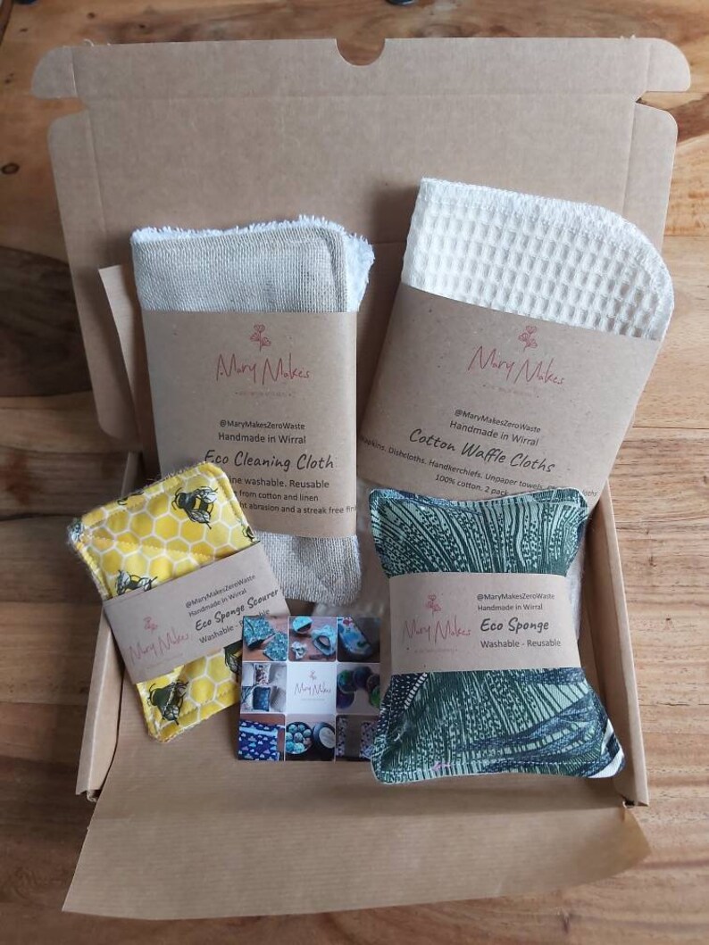 May include: A cardboard box filled with eco-friendly cleaning supplies. The box contains a set of cotton waffle cloths, a linen cleaning cloth, a yellow sponge with a bee design, and a green sponge with a leafy pattern. The products are labeled "Mary Makes Zero Waste" and "Handmade in Wirral".