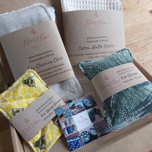 May include: A box filled with eco-friendly cleaning and kitchen products. The products include a waffle weave cotton cloth, a linen cleaning cloth, a yellow honeycomb sponge with bees, and a green eco-sponge. The products are handmade in Wirral and are reusable and washable.