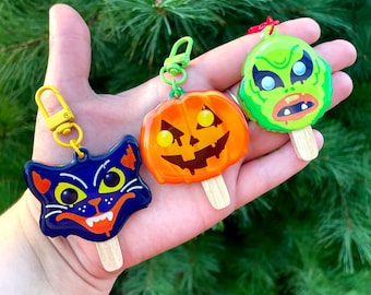 May include: Three Halloween-themed keychains shaped like popsicles. One is a dark blue cat face, one is an orange jack-o'-lantern, and one is a green monster face. Each features a small wooden stick at the bottom and a yellow or red metal clasp.