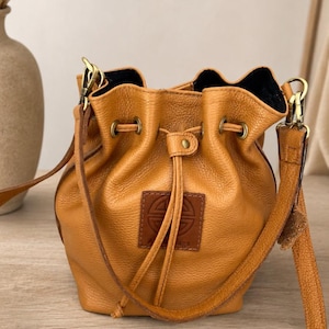 Può includere: Una borsa a secchiello in pelle color cuoio con chiusura a coulisse, finiture in metallo color ottone e una toppa quadrata in pelle con un logo circolare in rilievo sul davanti. La borsa include una tracolla regolabile con clip in metallo.