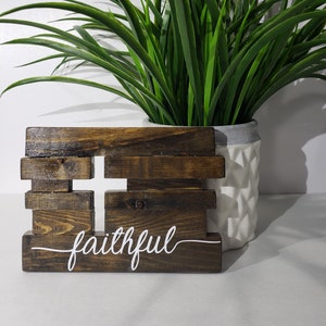 May include: A wooden cross sign with the word "faithful" written in white paint. The sign is made of rustic brown wood planks and is sitting on a white surface in front of a potted green plant.