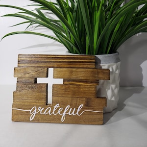 May include: A wooden sign with a white cross and the word "grateful" written in white cursive script. The sign is sitting on a white surface in front of a green plant.