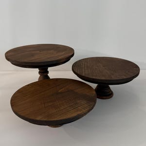 Puede incluir: Tres soportes circulares de madera para pasteles con un acabado te&ntilde;ido en marr&oacute;n oscuro. Cada soporte presenta una superficie superior redonda y plana y una base central de pedestal con un dise&ntilde;o torneado. La veta de la madera es visible.