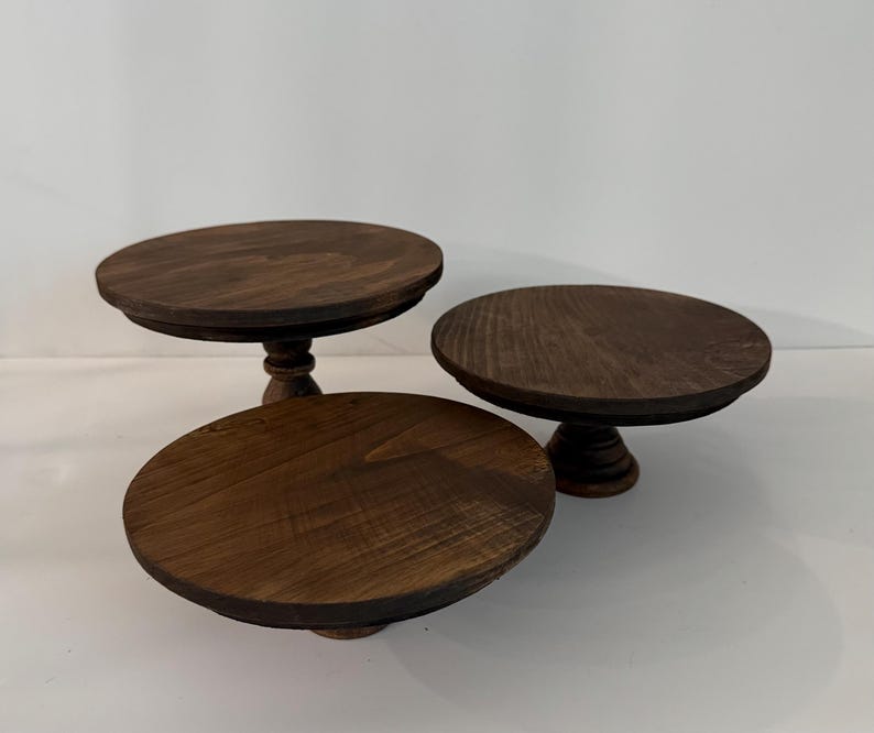 Puede incluir: Tres soportes circulares de madera para pasteles con un acabado te&ntilde;ido en marr&oacute;n oscuro. Cada soporte presenta una superficie superior redonda y plana y una base central de pedestal con un dise&ntilde;o torneado. La veta de la madera es visible.