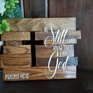 May include: A wooden sign with the words "Be Still and Know That I Am God" and the scripture reference "Psalms 46:10". The sign is made of reclaimed wood and has a rustic look.