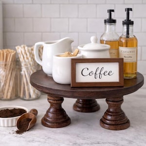 May include: A dark brown, three-legged wooden cake stand holds a white mug, creamer, sugar bowl, and a framed sign that reads "Coffee." Two glass bottles of syrup and a small bowl of coffee grounds are also present.