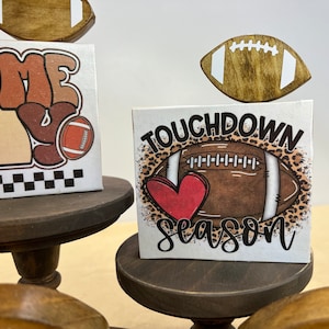 May include: Three tiered wooden stands with a football theme. The top tier has a brown football with white laces. The middle tier has a white sign with brown and orange text that says "Game Day" and a brown football with white laces. The bottom tier has a white sign with brown text that says "Touchdown Season" and a brown football with white laces and a red heart.