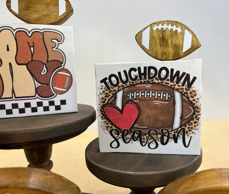 May include: Three tiered wooden stands with a football theme. The top tier has a brown football with white laces. The middle tier has a white sign with brown and orange text that says "Game Day" and a brown football with white laces. The bottom tier has a white sign with brown text that says "Touchdown Season" and a brown football with white laces and a red heart.