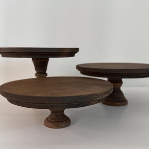 Puede incluir: Tres soportes para pasteles de madera de color marr&oacute;n oscuro de diferentes alturas, cada uno con una parte superior circular plana con borde biselado y una base de pedestal central. Los soportes est&aacute;n dispuestos en grupo.