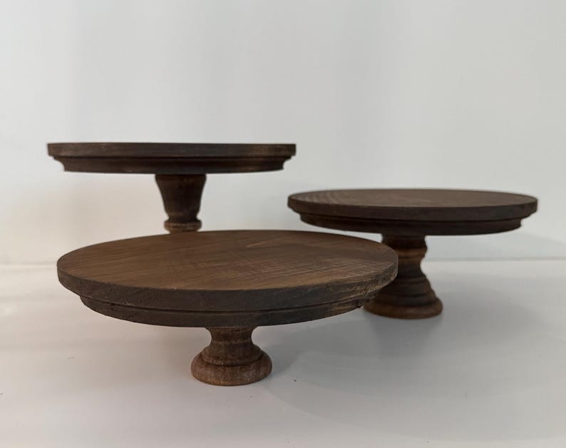 Puede incluir: Tres soportes para pasteles de madera de color marr&oacute;n oscuro de diferentes alturas, cada uno con una parte superior circular plana con borde biselado y una base de pedestal central. Los soportes est&aacute;n dispuestos en grupo.