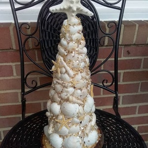 May include: A white seashell Christmas tree with a white starfish on top. The tree is made of various sizes and shapes of seashells and is sitting on a wooden base.