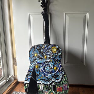 K&ouml;nnte beinhalten: Akustikgitarre mit einem Mosaikdesign, das eine Sternennachtszene darstellt. Das Mosaik weist Blaut&ouml;ne, Gelb- und Gr&uuml;nt&ouml;ne auf, mit einer schwarzen Bergsilhouette und bunten H&auml;usern am unteren Rand. Die Gitarre hat einen schwarzen Hals und einen schwarzen Gurt.