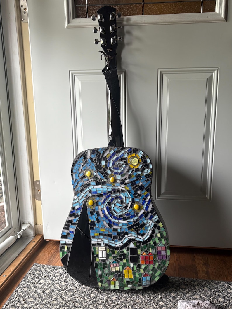 K&ouml;nnte beinhalten: Akustikgitarre mit einem Mosaikdesign, das eine Sternennachtszene darstellt. Das Mosaik weist Blaut&ouml;ne, Gelb- und Gr&uuml;nt&ouml;ne auf, mit einer schwarzen Bergsilhouette und bunten H&auml;usern am unteren Rand. Die Gitarre hat einen schwarzen Hals und einen schwarzen Gurt.