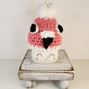 May include: A pink crocheted hat with a white pom pom and black eyes and beak sits on top of a white mug with a smiling face. The mug is on a white wooden stand with three legs.