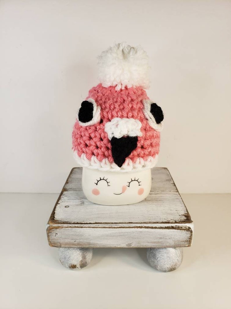 May include: A pink crocheted hat with a white pom pom and black eyes and beak sits on top of a white mug with a smiling face. The mug is on a white wooden stand with three legs.