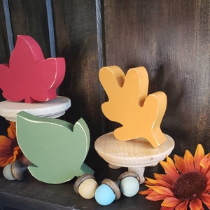 May include: Three wooden fall leaves on small wooden stands. The leaves are painted red, orange, and green. The stands have three legs each.
