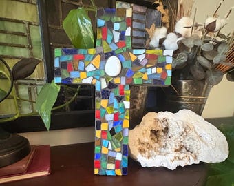 May include: A colorful mosaic cross with a white center, featuring a variety of glass tiles in shades of blue, green, red, and yellow. The cross is displayed on a dark wooden surface, with a geode and decorative items in the background.