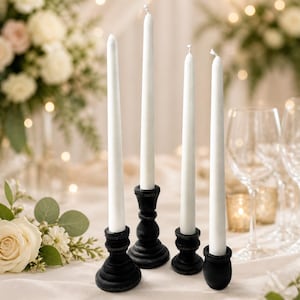 Peut inclure: Quatre bougies blanches effil&eacute;es dans des bougeoirs en bois noir. Les bougeoirs ont un design tourn&eacute; d&eacute;coratif. Les bougies sont dispos&eacute;es sur une nappe blanche, avec des compositions florales et de la verrerie en arri&egrave;re-plan.