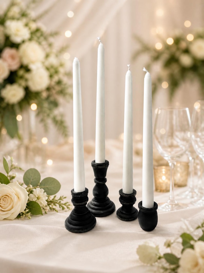 Peut inclure: Quatre bougies blanches effil&eacute;es dans des bougeoirs en bois noir. Les bougeoirs ont un design tourn&eacute; d&eacute;coratif. Les bougies sont dispos&eacute;es sur une nappe blanche, avec des compositions florales et de la verrerie en arri&egrave;re-plan.