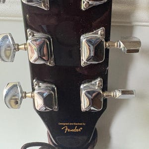 K&ouml;nnte beinhalten: Nahaufnahme der Kopfplatte einer dunkelbraunen Akustikgitarre. Die silbernen Stimmwirbel sind sichtbar, zusammen mit dem Text "Designed and Backed by Fender&reg;". Eine braune Gitarrensaite ist um die Kopfplatte gewickelt.