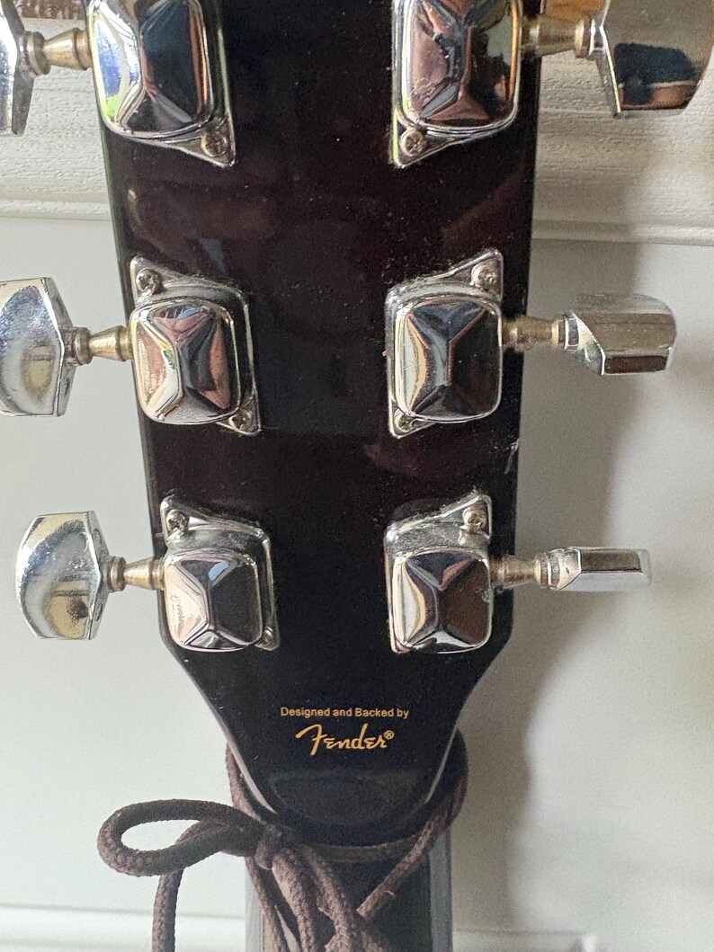 K&ouml;nnte beinhalten: Nahaufnahme der Kopfplatte einer dunkelbraunen Akustikgitarre. Die silbernen Stimmwirbel sind sichtbar, zusammen mit dem Text "Designed and Backed by Fender&reg;". Eine braune Gitarrensaite ist um die Kopfplatte gewickelt.