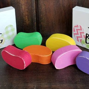May include: Two white wooden signs with the text "Happy Spring" and "Jelly Beans". The signs are decorated with colourful patterned paper. There are six colourful jelly bean shaped sweets in front of the signs.