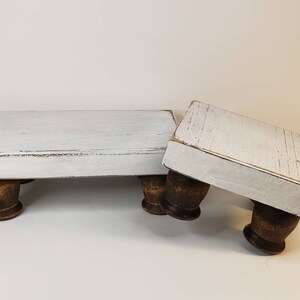 May include: Two white wooden tiered stands with distressed paint and brown wooden legs. The stands are stacked on top of each other.